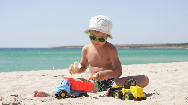 Child, boy playing with a plastic car in the sand on the beach and eating ice cream. Hot summer day on the beach.