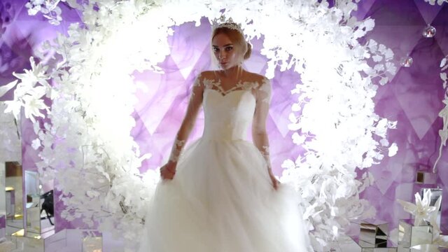 Beautiful young bride with a stylish hairstyle, a diadem on her head, earrings and a white dress whirls against the backdrop of a decorated arch and a purple background. Video