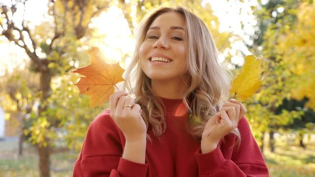 Attractive young blonde woman wearing red warm dress hides face with yellow maple leaves posing for camera in sunny autumn park closeup