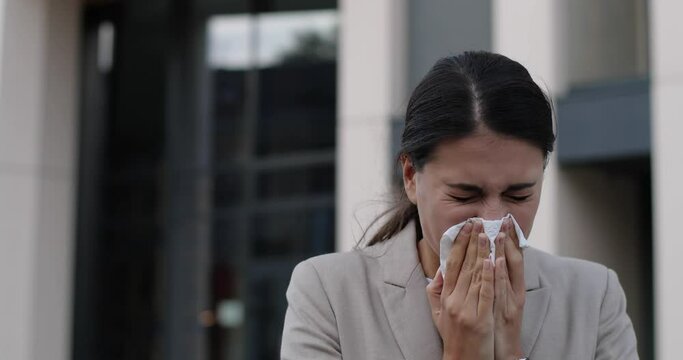 Close up of sick young female standing on street and coughing and sneezing. Portrait of unwell beautiful Caucasian woman blowing runny nose into tissue outdoors. Infection. Virus and disease concept