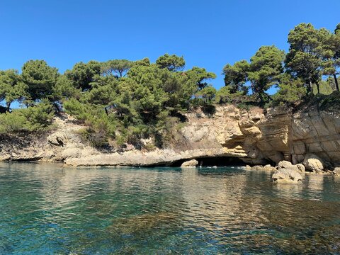 Rocky beach and turquoise calm rippled Adriatic sea surface,  scenic rocks and cave grotto, sunny summer day, green coniferous pine tree forest on the cliff