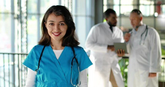 Portrait of Caucasian happy beautiful woman physician looking and smiling to camera and standing in clinic. Indoors. Pretty female doctor in hospital. Mixed-races doctors talking on background.