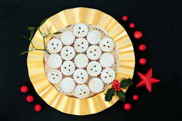 Traditional Christmas mince pie tarts on a gold plate with winter berry holly & mistletoe on black background with red bauble decorations. Festive food composition. Flat lay, top view, copy space.