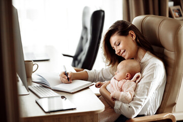 Happy young mother sit at the table, hold in arms little daughter, smiling, beautiful lady making notes, planning, write down ideas, business woman working at the home online