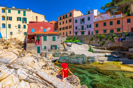 Woman in red dress sitting on cliffs at Marciana Marina looking famous houses of old village Borgo al Cotone in bay of Marciana Marina seaside. Elegant tourist on holiday travel on Elba Island, Italy.