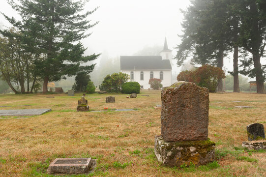 Headstone in a Small Cemetery Behind a White Wooden Church. Historic cemetery dating back to the 1800's situated behind a classic white church built in 1908. This foggy morning adds atmosphere.