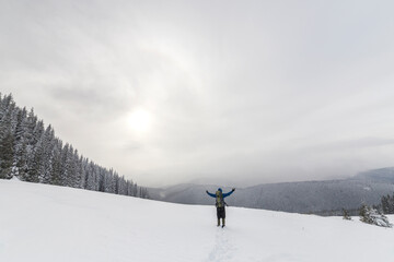 Back view of tourist hiker in warm clothing with backpack standing with raised arms on clearing covered with snow on spruce forest mountain and cloudy sky copy space background.
