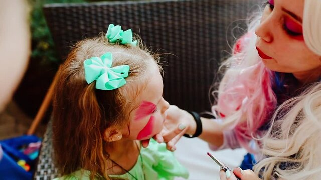Female animator making face painting for a young blond girl at the kid's party. Focus is at the girl. Slow motion video.