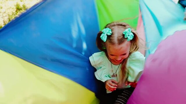Portrait of young pretty girl in green blouse and black pants with bows sitting inside the waving colourful toy parachute and smiling.