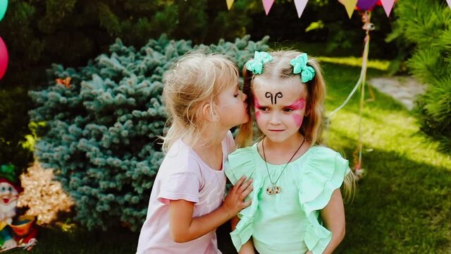 Two young blond girls with face painting kissing in the garden against party decoration. Slow motion video.