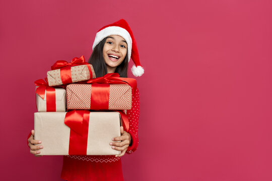 Excited funny indian latin kid child girl wears santa hat looking at camera holding many gifts boxes celebrating happy 2021 New Year isolated on red background. Merry Christmas presents shopping sale.