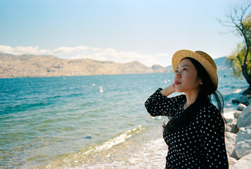 Asian Woman With A Black Polka Dot Dress and Straw Hat On Gazing Out At Okanagan Lake In British Columbia, Canada.