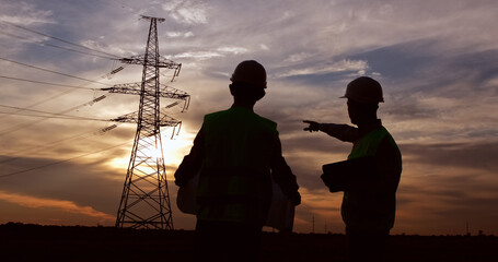 Two engineers in white safety helmets and green signal vests shake hands in front of a high-voltage pole at sunset.