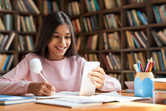 Indian Latin Child Preteen School Girl Holding Phone Distance Learning Class Using Mobile Application, Watching Online Lesson, Video Calling In App Making Notes Studying At Home, In Classroom.