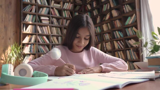 Cute smart indian latin preteen school girl pupil studying at home sitting at desk. Happy cute latin kid primary school student writing in exercise book doing homework, learning at table in classroom.