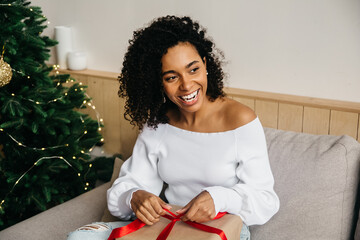 Happy laughing young woman opens box with christmas gift and looks away. Christmas lifestyle and black friday's concept. African american female model