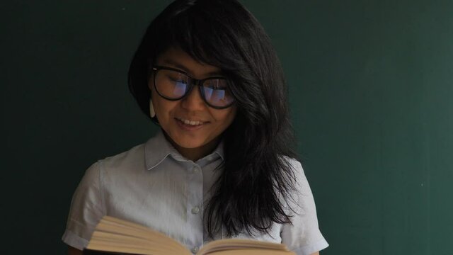 Emotional brunette with white earrings and glasses reads book and laughs standing near green wall at home slow motion close view