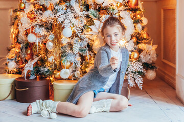 Fototapeta na wymiar Little girl with sparkler near Christmas tree on Christmas eve at home. Young kid in light bedroom with winter decoration Happy family at home Christmas New Year december time for celebration concept