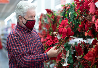 An elderly man with white hair choosing a Christmas garland in a store for the next holidays, wearing a red mask due to coronavirus infection