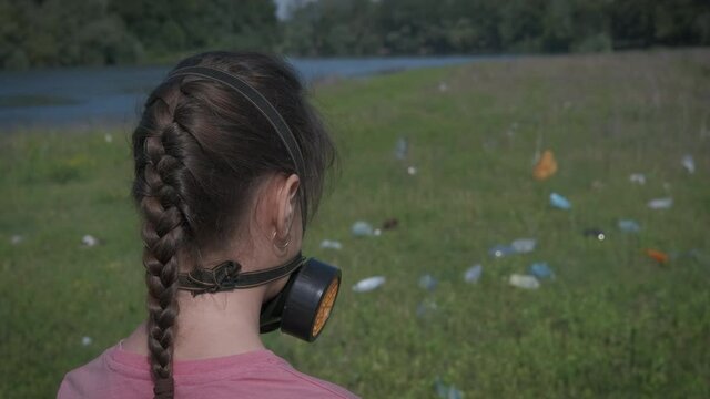 Environmental pollution. A girl in a respirator looks at the plastic garbage lying on the green lawn.