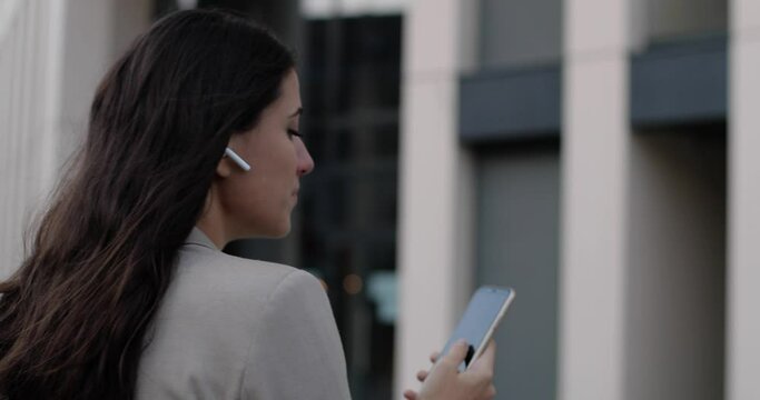 Rear of beautiful joyful young woman walking in city in wireless earphones and texting on smartphone while smiling. Close up of sick Caucasian female typing on cellphone outdoor. Health concept