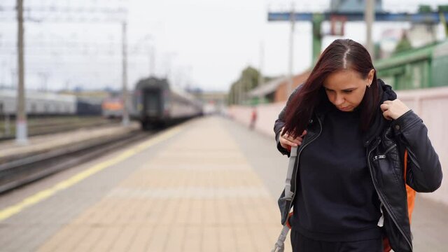 Young woman puts on backpack on her back, standing at train station. Adult female preparing for train ride. Concept of tourism, travel and recreation.