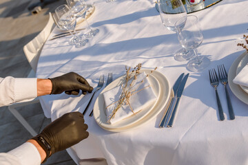 waiters hands in protective black gloves arrange a wedding party reception table decorated with flowers: plates, forks, knives and wine glasses.