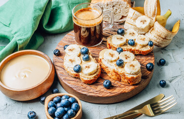 Buckwheat healthy bread with peanut butter, banana and blueberry on wooden board over concrete background. Selective focus. Summer breakfast.