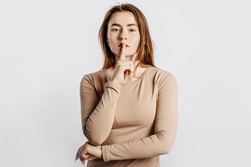 Shh gesture. Young beautiful serious girl holding a finger to her lips on a white isolated background. A woman asks to be silent, a place for advertising. Negative brunette in a beige jumper.