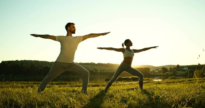 Caucasian female and male yogis standing at nature in morning and doing warrior asana. Sporty slim woman and man breathing, relaxing and doing yoga outdoor. Girl and guy starting day with training.