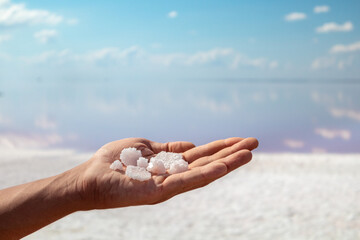 Hand holding bunch of pink white salt flakes crystals with pink salty lake and blue sky background. Spa resort sunny close-up