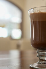 a vertically oriented shot of hot chocolate in a glass cup on a brown wooden table in a restaurant one bright sunny morning