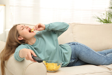 Lazy young woman with bowl of chips watching TV on sofa at home
