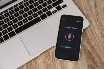 Smartphone with activated voice search app and laptop on wooden table