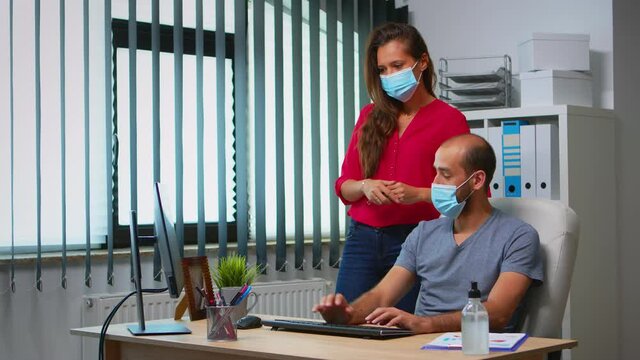 Workers talking wearing protection face masks in office room during coronavirus. Team in new normal office workspace in personal corporate company typing on computer keyboard and pointing at desktop