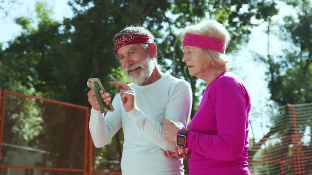 Caucasian mature grandparents using mobile phone fitness tracker application winning personal prizes celebrating reading good news training outside.