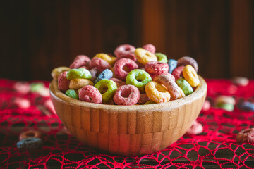 Colorful round fruit cereals in a wooden bowl on a red table, with some scattered outside the bowl. Wooden background behind with copy space. Breakfast ready.