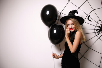 Naklejka premium Woman in witch hat with balloons posing near white wall decorated for Halloween