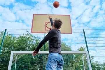 Teenager boy jumping with ball playing street basketball, back view