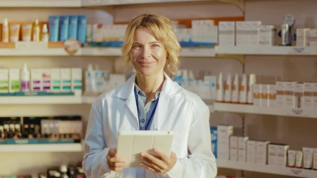 Portrait smiling woman in uniform pharmacist at pharmacy stand look at camera hold tablet computer. Diagnosis young pharmaceutics store job healthy medicine. Slow motion