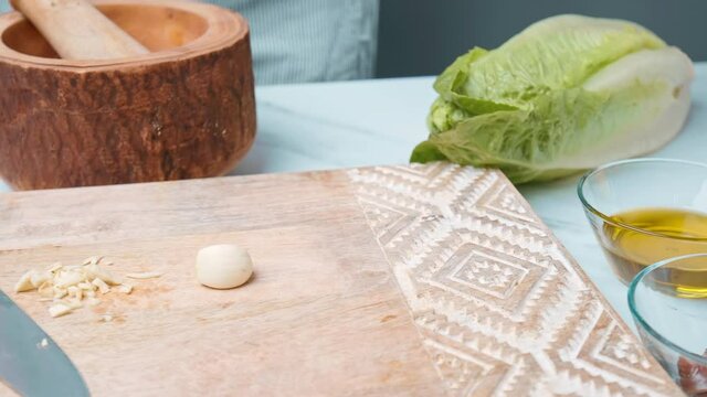 Chopping garlic on a wooden board for Caesar Salad dressing sauce in 4k. Close up of womans hands, finely cutting garlic and crashing it for a salad sauce.