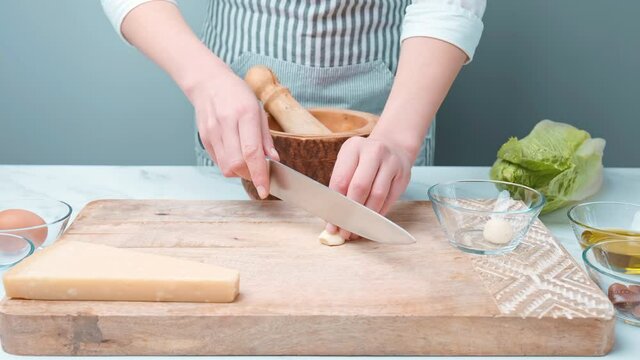 Chopping garlic on a wooden board for Caesar Salad dressing sauce in 4k. Close up of womans hands, finely cutting garlic and crashing it for a salad sauce.
