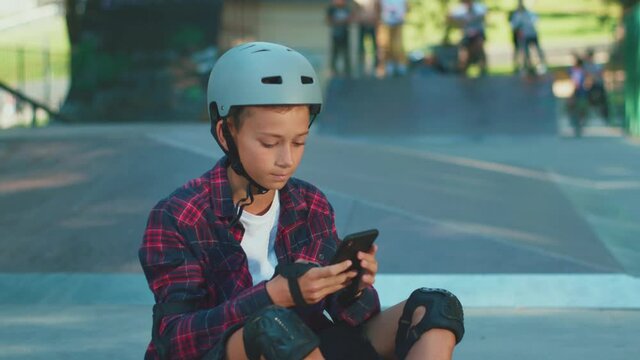 Teenager positive boy in helmet sits on floor of skatepark using smartphone taking selfie picture smiling happily. Skateboarding, sports, activity. Internet addiction.