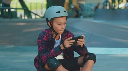 Young skateboarder with helmet using smartphone playing online game enjoying sitting on floor in skatepark. Hobby activities, smart children, internet addiction.