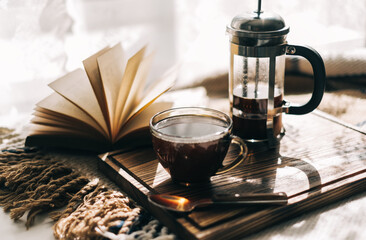 Coffee brewed in a French press and a cup on a wooden board with open book on a table. Morning at home, coziness and comfort.