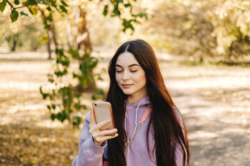 Beautiful young woman using smartphone and enjoying autumn weather in the park. Woman walking in the autumn park.