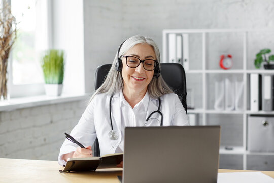 Portrait of senior grey-haired female doctor in her office using laptop for video chat with a patient. Online consultation with doctor for diagnoses and treatment recommendation. Telehealth concept.