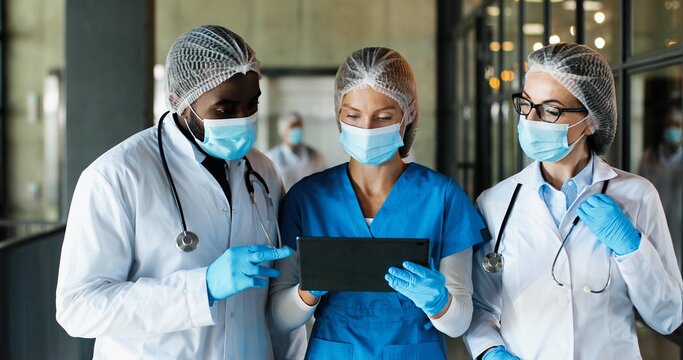 Mixed-races male and females doctors in hats, medical masks and gloves walking in hospital and talking, using tablet device. Multi ethnic man and women, medics discussing diagnosis. Corona concept.