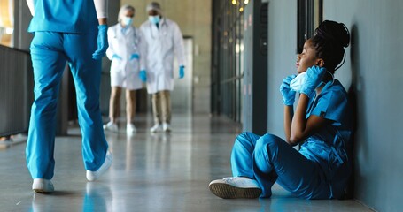 Tired sad African American woman doctor in uniform and gloves taking off medical mask and leaning on wall in despair. Upset female nurse resting, having break. Coronavirus. Rest at work. Covid-19.