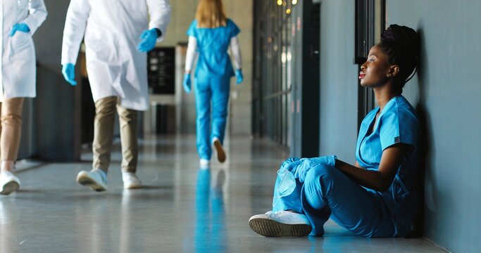 Tired Sad African American Woman Doctor In Uniform And Gloves Taking Off Medical Mask And Leaning On Wall In Despair. Upset Female Nurse Resting, Having Break. Coronavirus. Rest At Work. Covid-19.